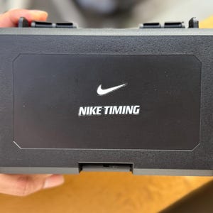 May include: A black rectangular case with the Nike swoosh logo and the words "NIKE TIMING" in white. The case has a textured surface and a hinged lid with two black latches.