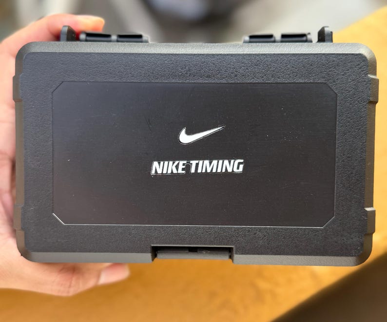 May include: A black rectangular case with the Nike swoosh logo and the words "NIKE TIMING" in white. The case has a textured surface and a hinged lid with two black latches.