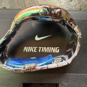 May include: A silver-toned Nike Timing watch with a stainless steel band. The watch face is not visible, but the watch has two buttons on the side. The watch is resting on a black surface with the Nike logo and the words "NIKE TIMING".