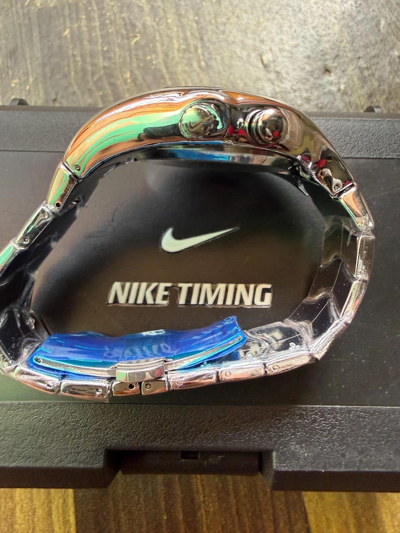 May include: A silver-toned Nike Timing watch with a stainless steel band. The watch face is not visible, but the watch has two buttons on the side. The watch is resting on a black surface with the Nike logo and the words "NIKE TIMING".