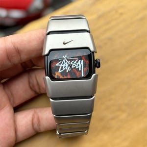 Könnte beinhalten: Eine silberfarbene Armbanduhr mit einem Metallgliederarmband und einem rechteckigen Zifferblatt. Das Zifferblatt zeigt ein braun-schwarzes Leopardenmuster mit dem weißen Schriftzug Stussy und einem kleinen schwarzen Nike-Logo.