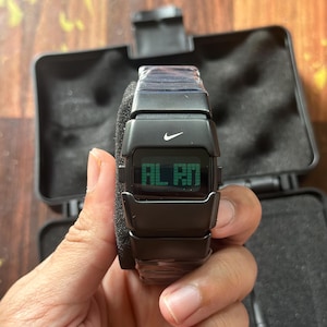 May include: Black digital watch with the Nike logo and the word "ALARM" displayed on the screen. The watch is in a black case with a foam insert. The watch is being held by a person.