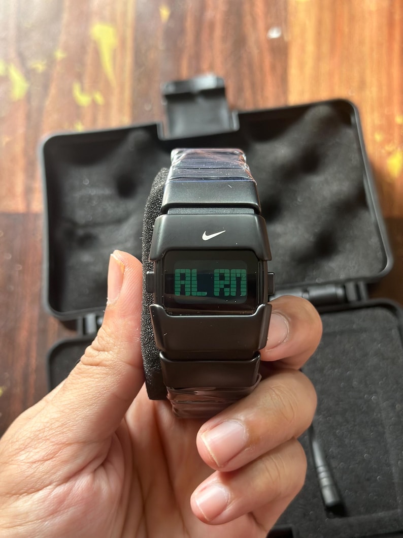 May include: Black digital watch with the Nike logo and the word "ALARM" displayed on the screen. The watch is in a black case with a foam insert. The watch is being held by a person.