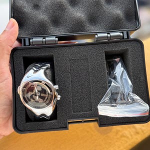 May include: A silver-toned sports watch with a black band, displayed in a black case with foam inserts. The watch face has a circular design with multiple dials and the word "SET". An additional accessory is included.