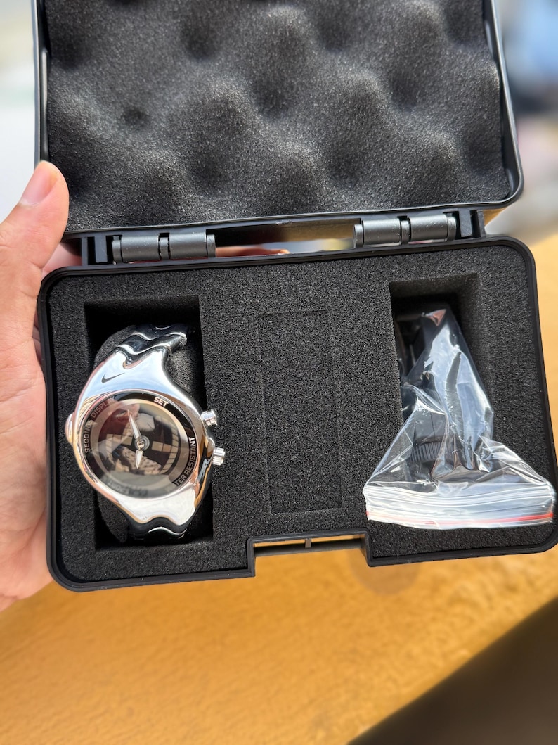May include: A silver-toned sports watch with a black band, displayed in a black case with foam inserts. The watch face has a circular design with multiple dials and the word "SET". An additional accessory is included.