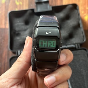 May include: Black digital watch with the Nike logo. The watch displays "CH RD" on a green screen. The watch is held in a hand and is in a black case.