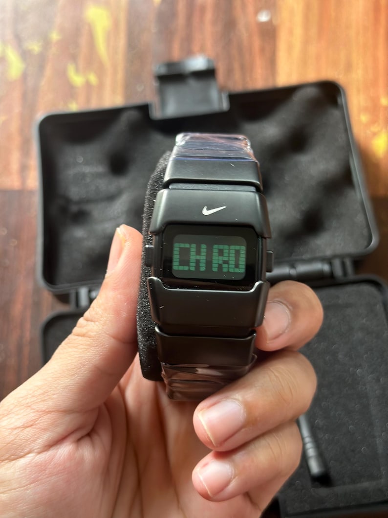 May include: Black digital watch with the Nike logo. The watch displays "CH RD" on a green screen. The watch is held in a hand and is in a black case.