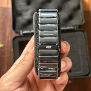 May include: A black Nike watch with a rectangular, segmented band. The watch is displayed in an open black case with a foam insert. The Nike logo is visible on the watch face.
