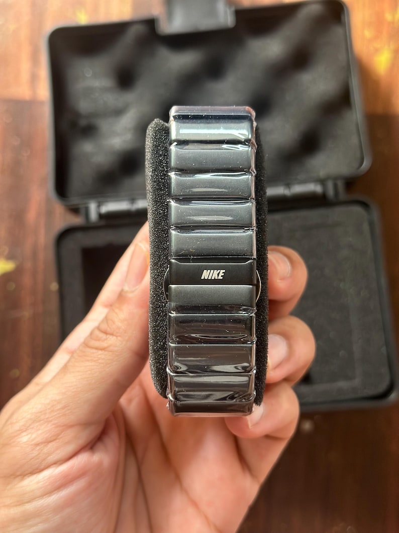 May include: A black Nike watch with a rectangular, segmented band. The watch is displayed in an open black case with a foam insert. The Nike logo is visible on the watch face.