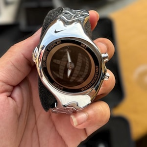 May include: A silver-toned Nike sports watch with a circular face. The watch features a black dial with a compass design and the words "SECONDS DISPLAY", "SPORT", and "WATER RESISTANT". The watch is held in a person's hand.