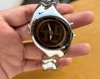May include: A silver-toned Nike sport watch with a unique wave-like design. The watch face displays a compass-like design with the words "SECONDS DISPLAY", "SPORT", "WATER RESISTANT", and "SET". The watch has a sleek, modern aesthetic.