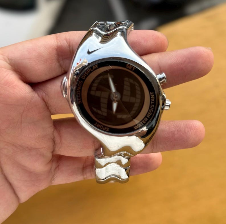 May include: A silver-toned Nike sport watch with a unique wave-like design. The watch face displays a compass-like design with the words "SECONDS DISPLAY", "SPORT", "WATER RESISTANT", and "SET". The watch has a sleek, modern aesthetic.