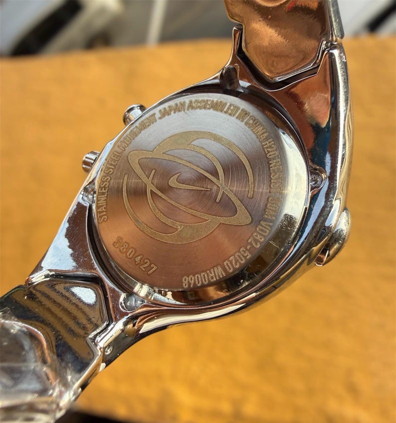 May include: A silver-toned Nike wristwatch, viewed from the back, showcasing the watch's stainless steel construction. The watch face features the Nike logo and the text "Stainless Steel Movement Japan Assembled in China H20 Resist 50M".