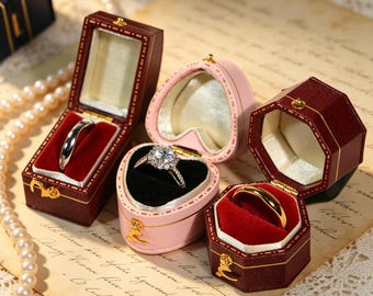 May include: An assortment of open ring boxes in various shapes and colors. The boxes are burgundy, pink, and heart-shaped, with gold accents and red or black velvet interiors. Each box holds a different ring, including a silver band and a diamond engagement ring.