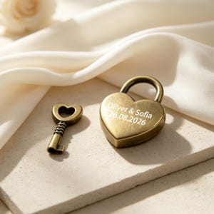 May include: A heart-shaped brass padlock and a matching key. The padlock is engraved with the text Oliver & Sofia and the date 26.08.2026. Both items feature a brushed metallic finish and are positioned together on a flat, light-colored surface.