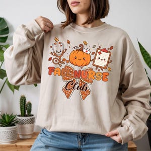 May include: A beige crewneck sweatshirt featuring a graphic print of a smiling pumpkin wearing a nurse cap, a stethoscope, and a clipboard. Below the characters, the text reads "FALL NURSE CLUB" in orange letters over a large patterned bow.