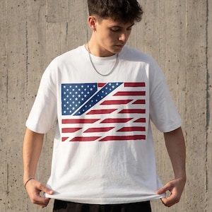 Könnte beinhalten: Weißes T-Shirt mit einem stilisierten Design der amerikanischen Flagge. Die Flaggen-Grafik zeigt einen blauen Bereich mit weißen Sternen und rot-weißen Streifen. Das Shirt ist ein lässiger Stil mit kurzen Ärmeln.