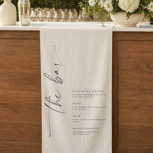 May include: A cream-coloured fabric table runner featuring the black printed text The bar, followed by a menu listing signature drinks, wine, beer, and non-alcoholic options. The runner has a textured weave and hangs vertically over a wooden bar.