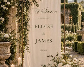 May include: A tall, rectangular off-white fabric wedding welcome sign suspended on a bronze metal frame. The sign features black text reading Welcome to the wedding of Eloise & James 07.27.2027, adorned with white floral arrangements.