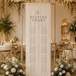 May include: A tall, rectangular, light-colored fabric seating chart banner suspended from a thin gold metal frame. The banner features the text "the SEATING CHART" at the top, followed by a list of names organized into ten numbered tables.