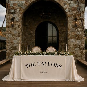 Puede incluir: Una mesa de boda rectangular cubierta con un mantel color crema que presenta un camino de mesa central con el texto negro impreso THE TAYLORS y EST. 2026. La mesa está decorada con arreglos florales blancos y seis velas altas.