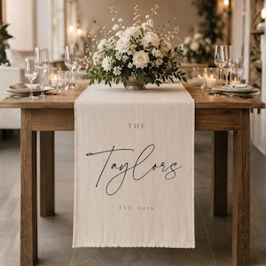 May include: A cream-coloured fabric table runner with frayed edges, featuring the printed text THE Taylors EST. 2026 in black ink. The runner is draped over a wooden table, positioned beneath a floral centrepiece with white roses and greenery.