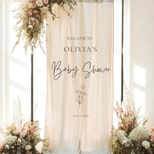Op de afbeelding: Een beige stoffen banner hangend aan een zwarte metalen standaard, met de tekst WELCOME TO OLIVIA'S Baby Shower en JULY 20, 2026, en een lijntekening van bloemen. De banner is versierd met arrangementen van witte en roze bloemen.
