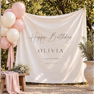 Op de afbeelding: Een crèmekleurige stoffen achtergrond met de tekst Happy Birthday, OLIVIA en 05.05.2027. Het geheel is versierd met een tros lichtroze en witte ballonnen vastgebonden met een roze lint, naast een houten bankje en een stenen plantenbak.