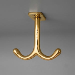 May include: A polished gold-tone metal ceiling hook featuring a central vertical post that branches into two curved arms. The base is a rectangular plate with two visible screw holes for mounting, providing a dual-pronged hanging solution.
