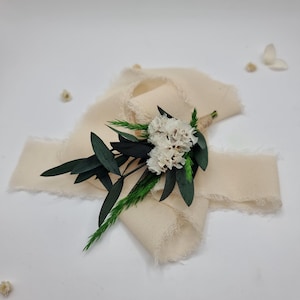 Könnte beinhalten: Ein cremefarbenes Stoffband mit ausgefransten Kanten, verziert mit einem kleinen Blumenarrangement. Das Arrangement besteht aus weißen Blüten, dunkelgrünen Blättern und grünen Zweigen. Die Artikel sind auf einem weißen Hintergrund angeordnet.