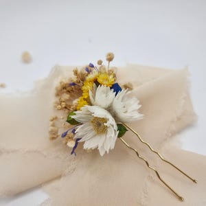 May include: A floral hair pin with white, yellow, and blue dried flowers. The arrangement includes small, dried blooms and green leaves, secured with gold-toned bobby pins. The flowers rest on a light beige fabric.
