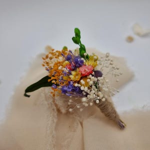 Könnte beinhalten: Ein farbenfrohes Gesteck mit Trockenblumen in Orange, Lila, Gelb und Rosa, akzentuiert mit weißem Schleierkraut. Die Stiele sind mit Bindfaden umwickelt und liegen auf einem cremefarbenen Stoff.