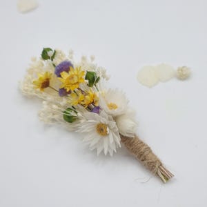 Könnte beinhalten: Eine handgefertigte Ansteckblume mit getrockneten Blumen in Weiß, Gelb und Lila. Das Arrangement umfasst gänseblümchenartige Blüten und zarte Füllblumen, mit einem mit Bindfaden umwickelten Stiel. Ideal für Hochzeiten oder besondere Anlässe.