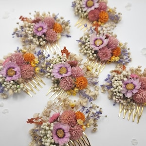 Op de afbeelding: Een verzameling van zeven haarkammen, elk met een goudkleurige metalen basis versierd met een arrangement van gedroogde bloemen, waaronder roze madeliefjes, oranje kogelamarant, wit gipskruid en kleine takjes paarse lavendel.