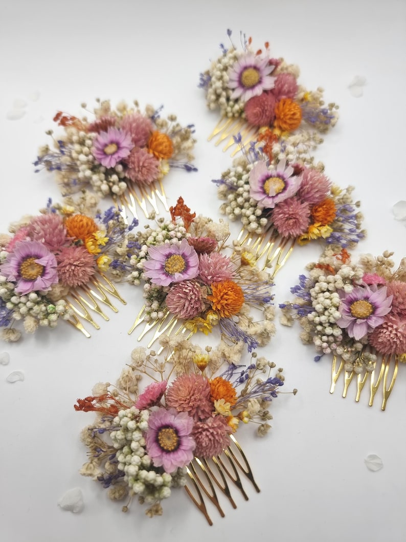Op de afbeelding: Een verzameling van zeven haarkammen, elk met een goudkleurige metalen basis versierd met een arrangement van gedroogde bloemen, waaronder roze madeliefjes, oranje kogelamarant, wit gipskruid en kleine takjes paarse lavendel.