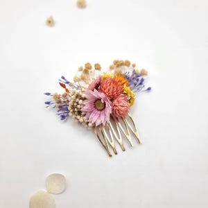 Op de afbeelding: Een haarkam van goudkleurig metaal versierd met een arrangement van gedroogde bloemen in roze, oranje, gele en paarse tinten, inclusief wit gipskruid en een centrale roze bloem, met losse bloemblaadjes verspreid in de nabijheid.