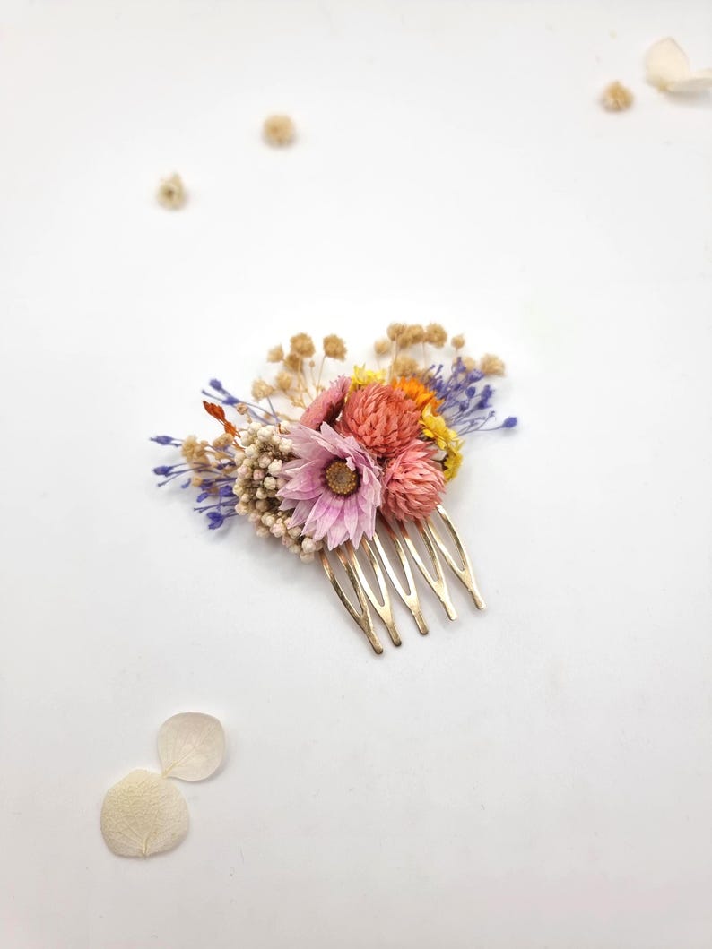 Op de afbeelding: Een haarkam van goudkleurig metaal versierd met een arrangement van gedroogde bloemen in roze, oranje, gele en paarse tinten, inclusief wit gipskruid en een centrale roze bloem, met losse bloemblaadjes verspreid in de nabijheid.