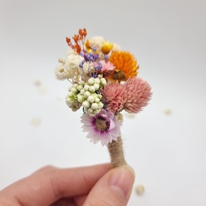 Op de afbeelding: Een kleine corsage samengesteld uit gedroogde bloemen in tinten oranje, roze, wit en paars. Het arrangement bevat kleine trossen witte bessen, oranje strobloemen, roze klaverachtige bloemen en een enkel lichtpaars madeliefje.