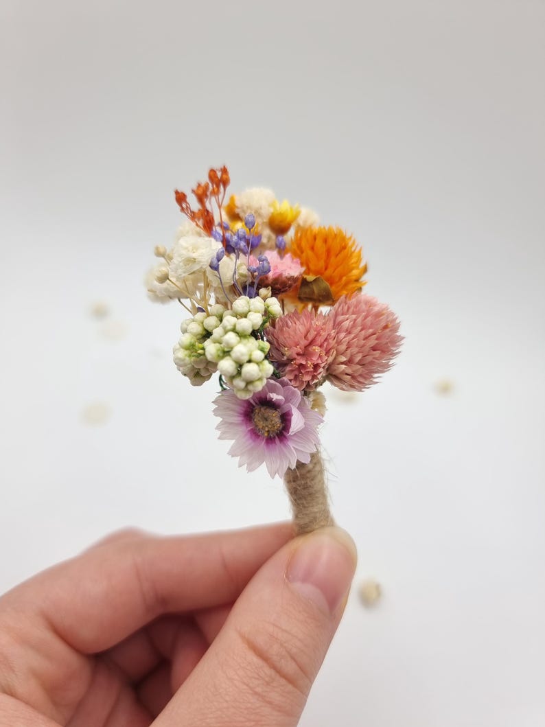Op de afbeelding: Een kleine corsage samengesteld uit gedroogde bloemen in tinten oranje, roze, wit en paars. Het arrangement bevat kleine trossen witte bessen, oranje strobloemen, roze klaverachtige bloemen en een enkel lichtpaars madeliefje.