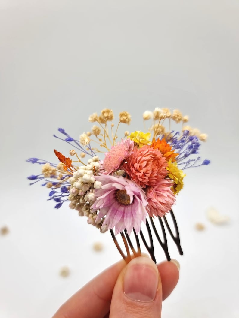 Op de afbeelding: Een haarkam met een boeketje gedroogde bloemen in roze, oranje, gele en blauwe tinten, geaccentueerd met kleine witte en beige takjes. Het decoratieve arrangement is bevestigd op een donkerbruine, meertandige haarkam van kunststof.