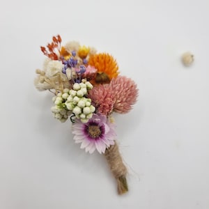 Op de afbeelding: Een corsage bestaande uit een boeketje gedroogde bloemen met een lichtpaarse margriet, roze klaverachtige bloemen, oranje strobloemen, witte bessen en kleine takjes blauwe en rode vulbloemen, met de stelen omwikkeld met natuurlijk touw.
