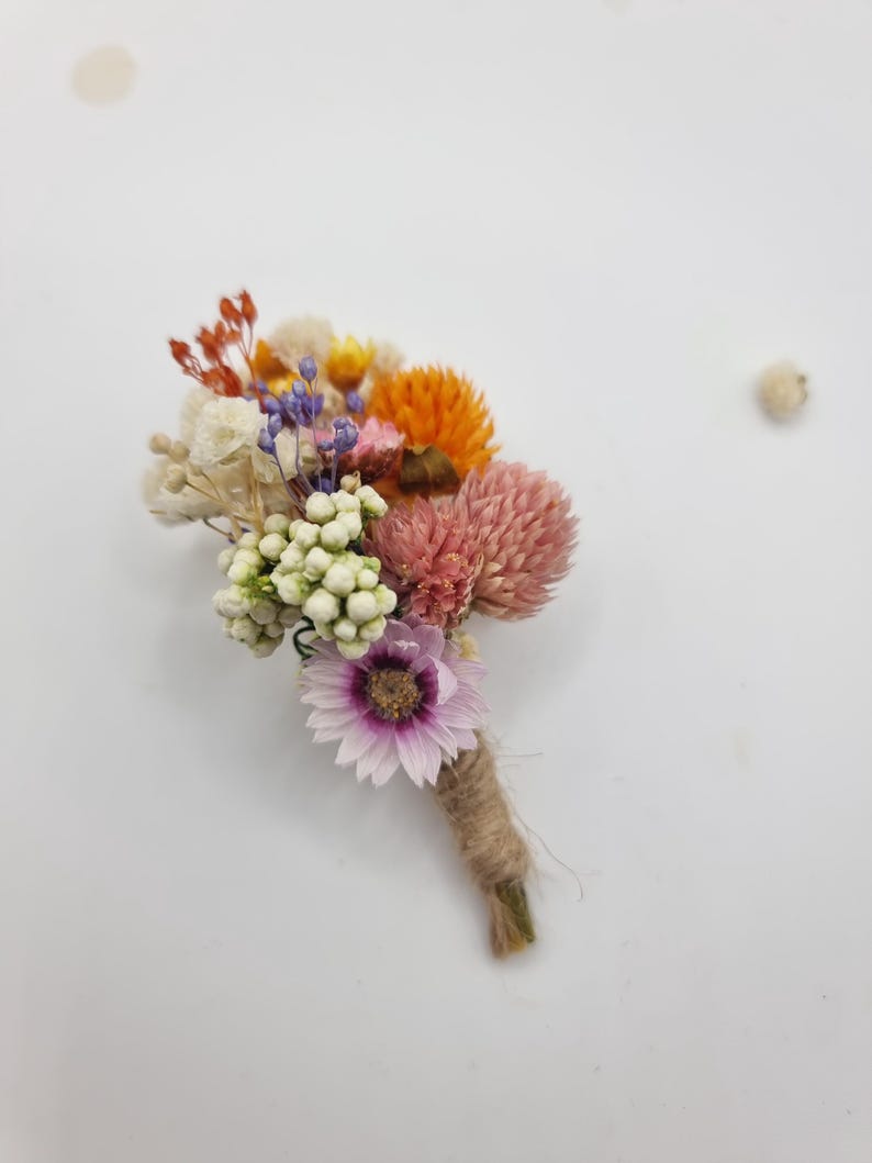 Op de afbeelding: Een corsage bestaande uit een boeketje gedroogde bloemen met een lichtpaarse margriet, roze klaverachtige bloemen, oranje strobloemen, witte bessen en kleine takjes blauwe en rode vulbloemen, met de stelen omwikkeld met natuurlijk touw.