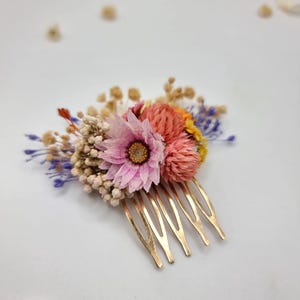 Op de afbeelding: Een goudkleurige haarkam versierd met een arrangement van gedroogde bloemen, waaronder een opvallende roze bloem, ronde koraalkleurige bloesems, kleine trosjes witte gipskruid en delicate takjes paarse en gele gedroogde flora.