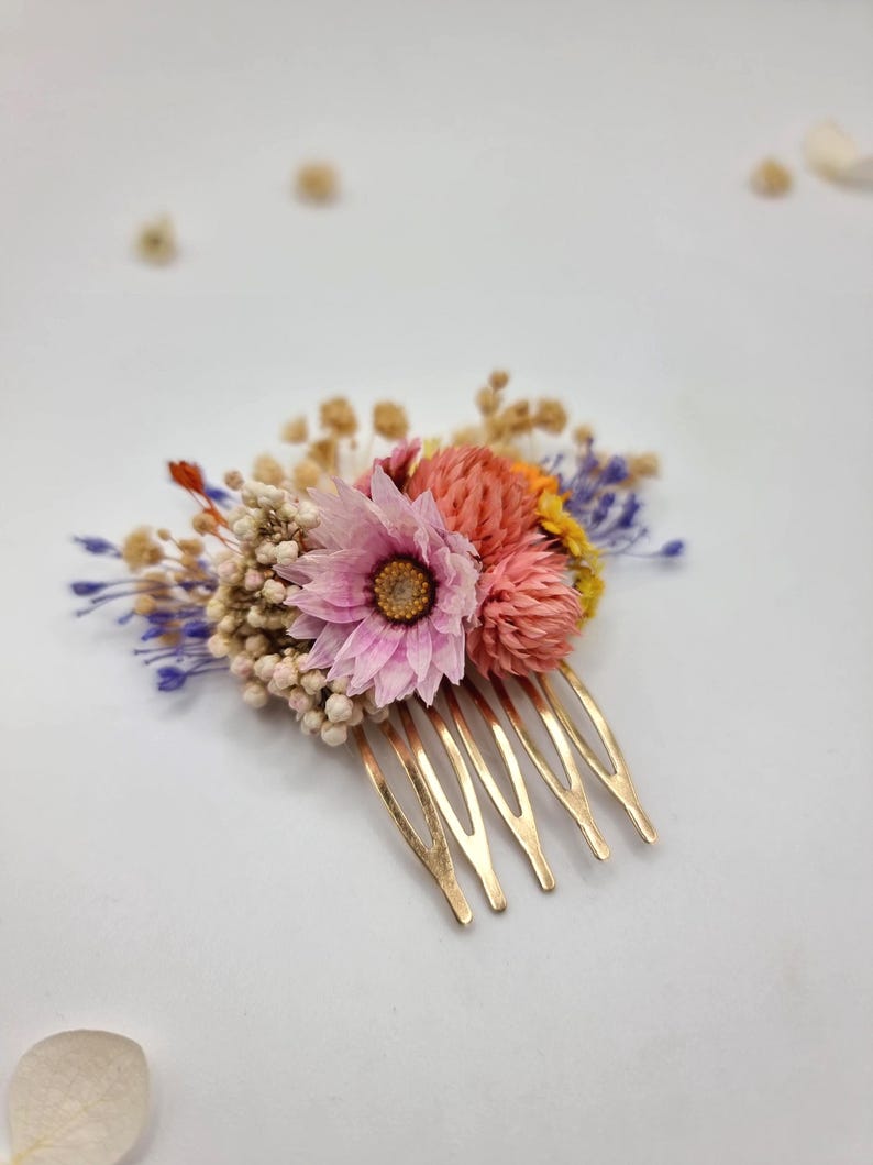 Op de afbeelding: Een goudkleurige haarkam versierd met een arrangement van gedroogde bloemen, waaronder een opvallende roze bloem, ronde koraalkleurige bloesems, kleine trosjes witte gipskruid en delicate takjes paarse en gele gedroogde flora.
