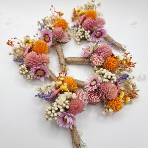 Op de afbeelding: Een set van drie corsages met gedroogde bloemen in de kleuren roze, oranje, geel en wit. Elk arrangement is omwikkeld met natuurlijk jutegaren en is voorzien van kleine spelden met een parelkop aan de onderkant van de stelen.