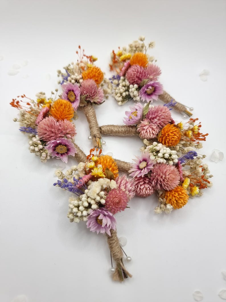 Op de afbeelding: Een set van drie corsages met gedroogde bloemen in de kleuren roze, oranje, geel en wit. Elk arrangement is omwikkeld met natuurlijk jutegaren en is voorzien van kleine spelden met een parelkop aan de onderkant van de stelen.