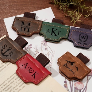 May include: A selection of personalised leather pen holders in various colours, including brown, green, and purple. Each holder features custom initials, names, or a mountain graphic, and an elastic loop for securing to a notebook or journal.
