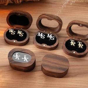 Peut inclure: Plusieurs boîtes à bijoux en bois, certaines ouvertes, affichant des boucles d'oreilles initiales en or et en argent. Les boîtes sont ovales avec une finition en bois foncé et des intérieurs en velours noir. Les boucles d'oreilles présentent les initiales WS, MS, TR et JK.