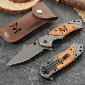 Peut inclure: Un couteau pliant avec une lame et un manche en métal gris, doté d'une incrustation en bois clair gravée du nom Michael et des initiales MVI. Un étui en cuir marron assorti et un couteau fermé avec un clip de poche noir sont inclus.