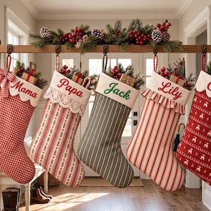 May include: Five hanging Christmas stockings with white cuffs embroidered with the names Mama, Papa, Jack, Lily, and Coco. The stockings feature various patterns including red gingham, striped, and reindeer prints, each filled with greenery and small gift boxes.